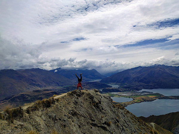 Man doing a handstand on a mountain ridge in front of a spectacular mountain & lake landscape
