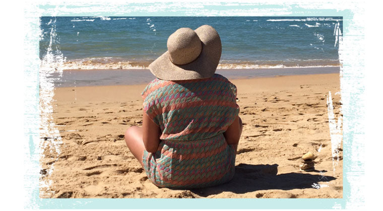 Claire wearing a sunhat & sitting on a sandy beach watching the waves on a sunny day