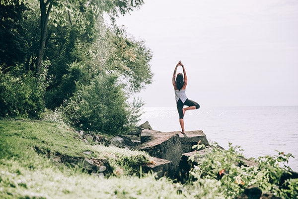 Young woman practicing yoga on a rock in front of the sea, surrounded by green grass, bushes & trees